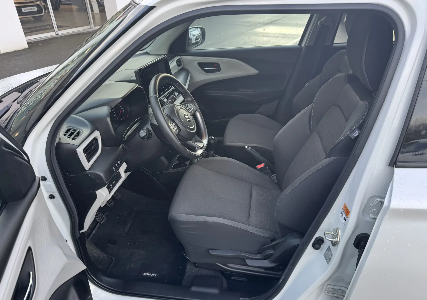 Intérieur de la Suzuki Swift 1.2 Privilège blanc, vue côté gauche montrant sièges noirs et tableau de bord moderne.