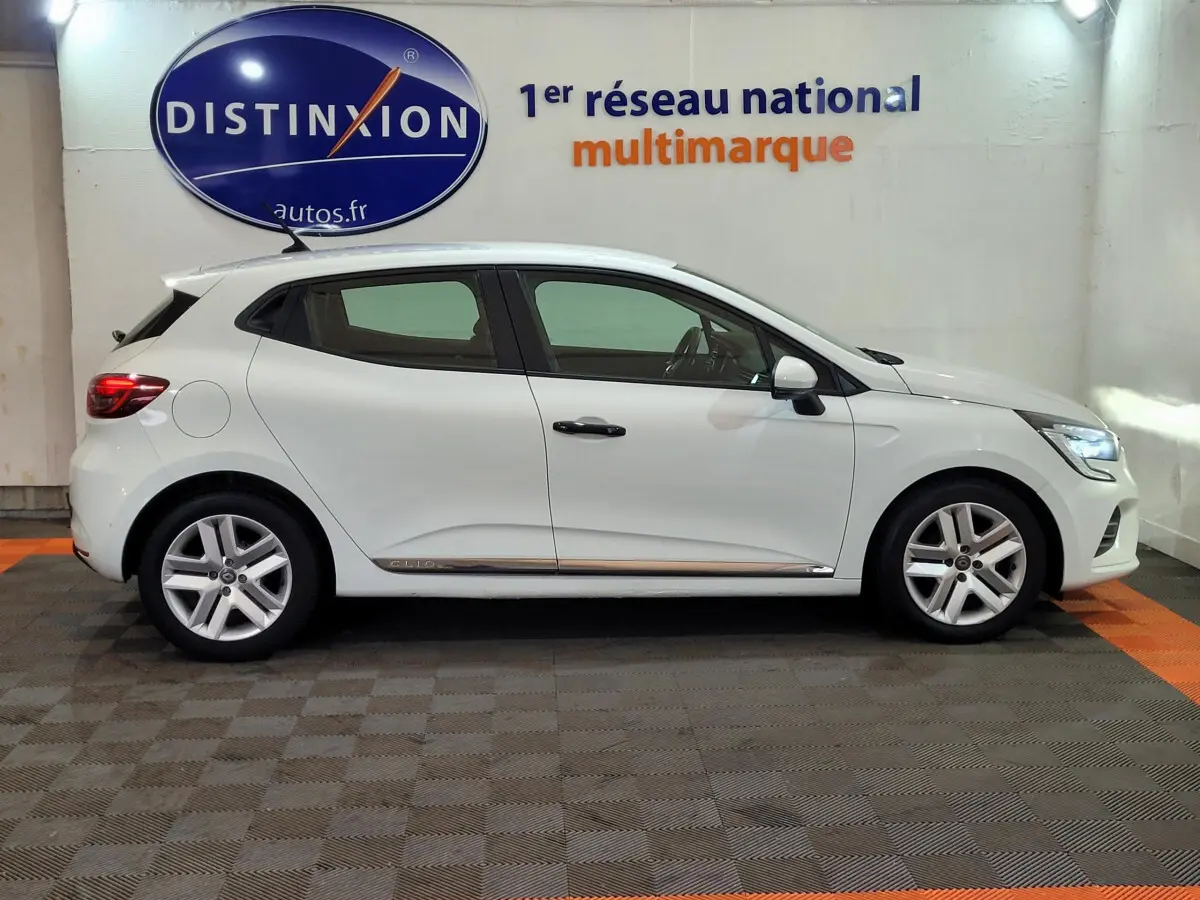Profil côté gauche d'une Renault Clio Business blanche 2021, avec jantes alliage et feux arrière allumés, en intérieur showroom.