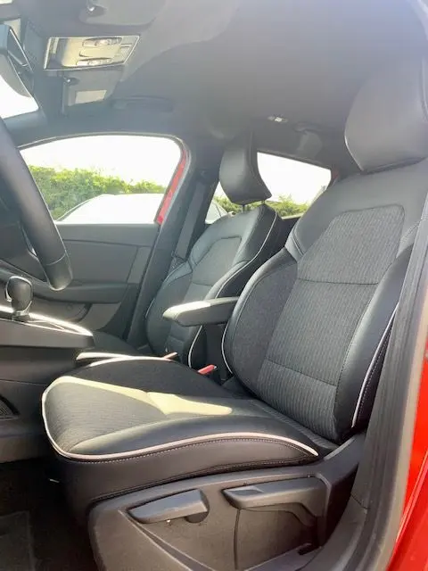 Vue intérieure côté conducteur de la Renault Clio V rouge 2021, mettant en valeur les sièges tissu et l'accoudoir central.