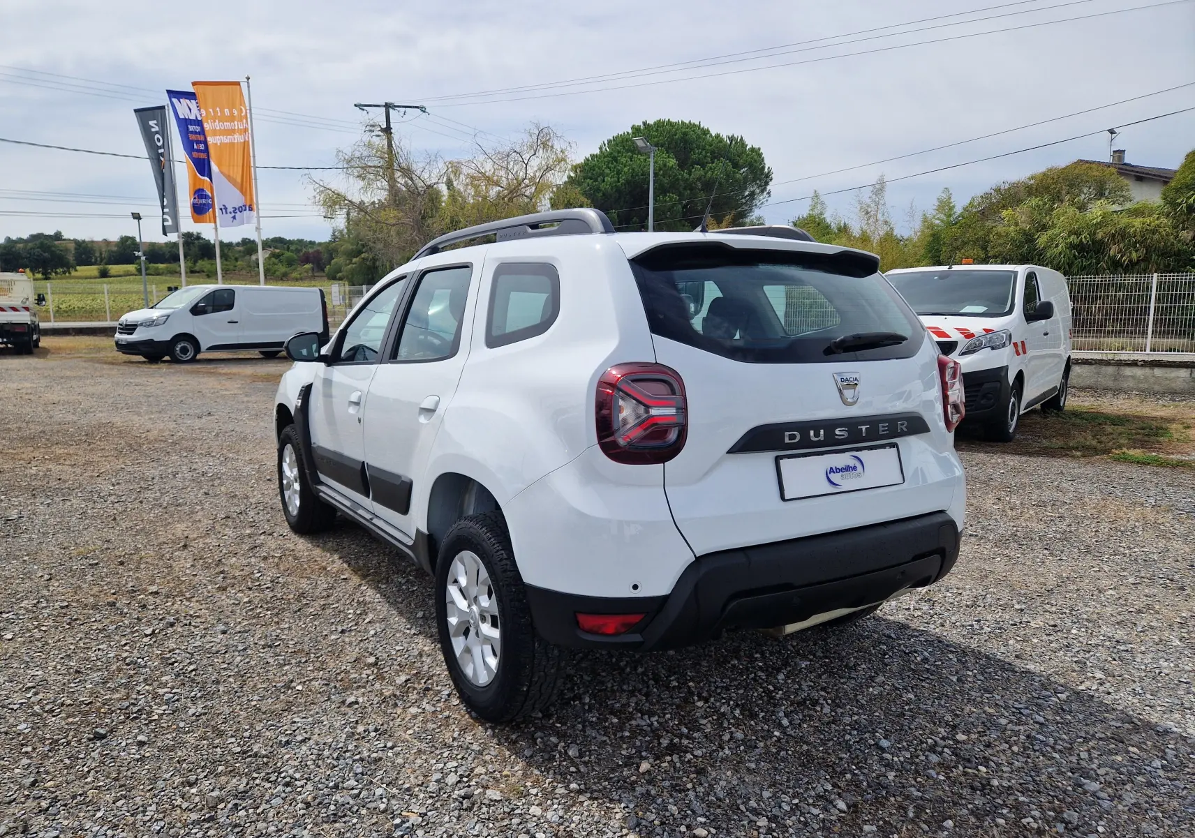 Vue 3/4 arrière droite d'un Dacia Duster blanc 2022 avec barres de toit et protections noires sur un sol gravillonné.