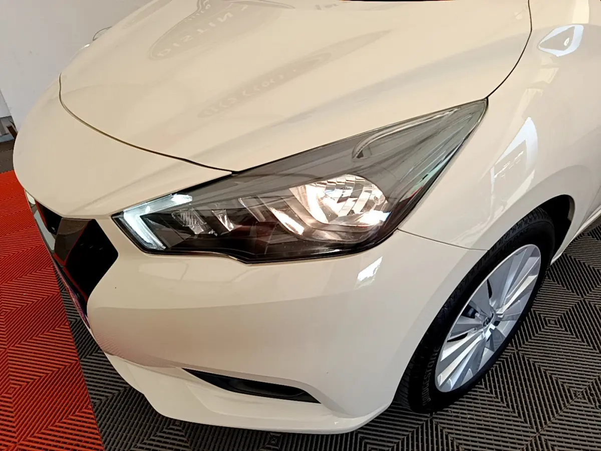 Vue 3/4 avant droit d'une Nissan Micra IG-T 92 Acenta blanche avec phares allumés et jantes alliage visibles.