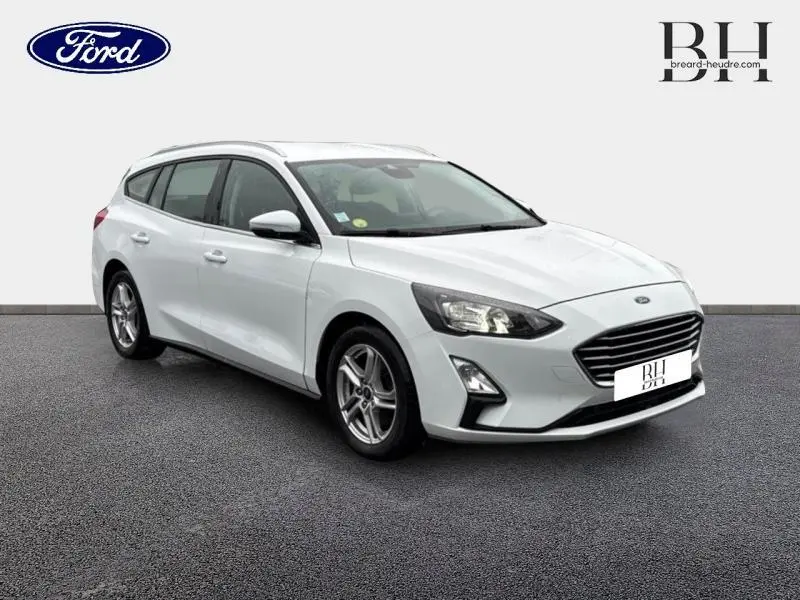 Ford Focus SW blanc glacier vue 3/4 avant droit avec jantes alu et barres de toit visibles.