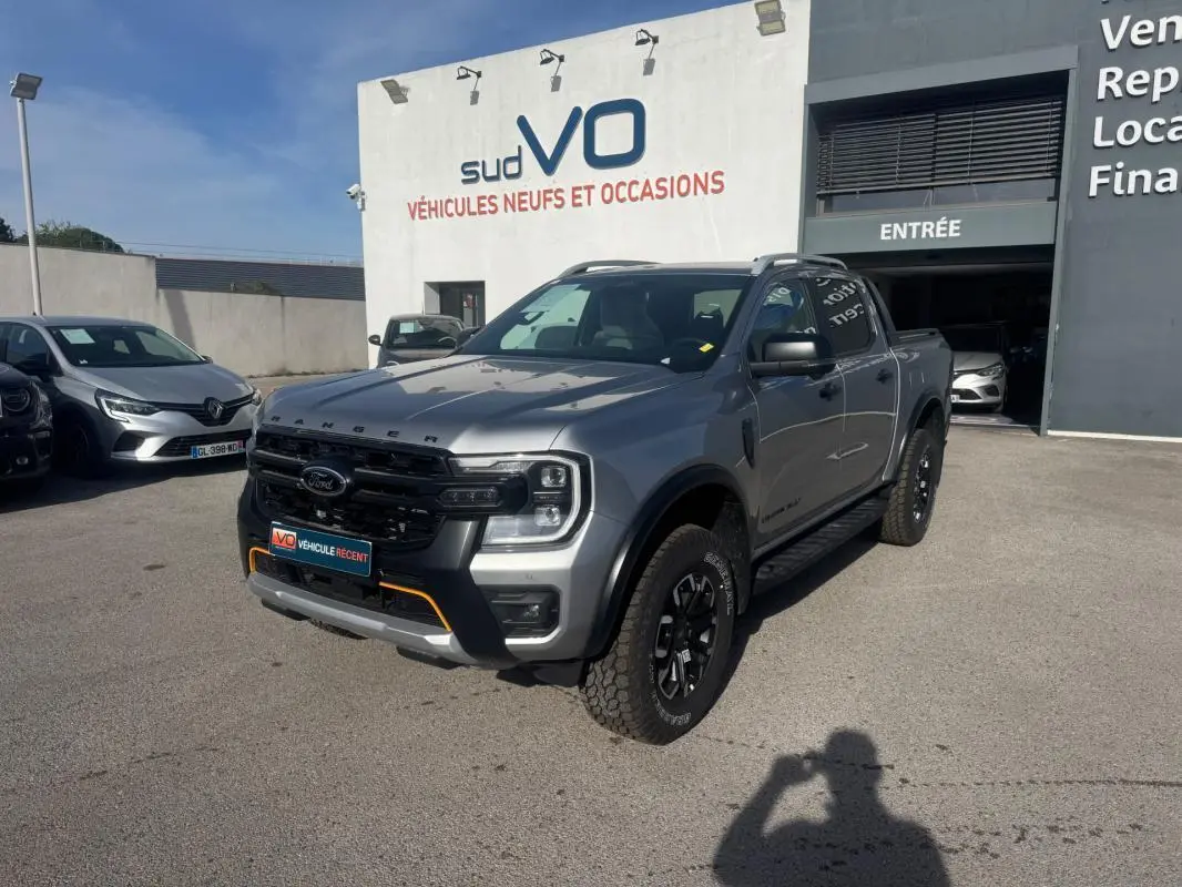 Ford Ranger Wildtrak gris en 3/4 avant droit, avec barres de toit et jantes alliage noires visibles.