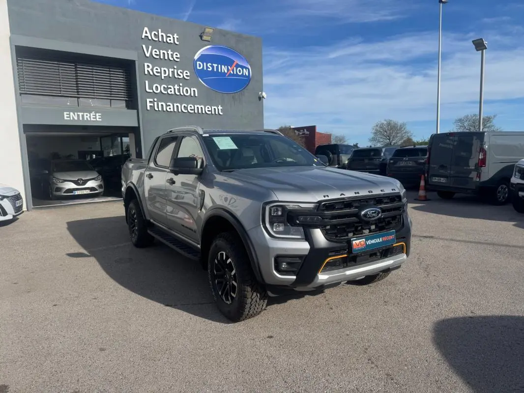 Ford Ranger gris en 3/4 avant droit, avec jantes alliage noires et barres de toit visibles, stationné devant un garage.