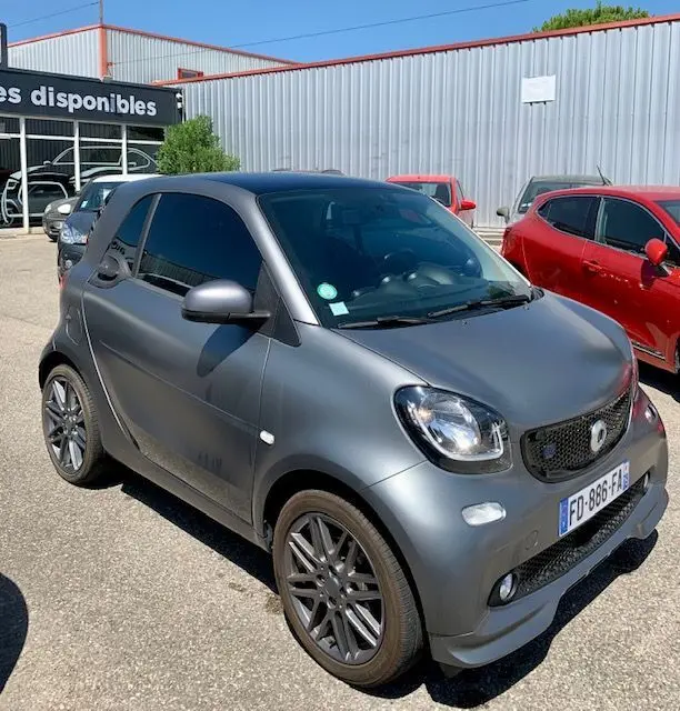 Vue 3/4 avant droite d'une Smart Fortwo Coupé électrique gris mat avec jantes Brabus style sur parking extérieur.
