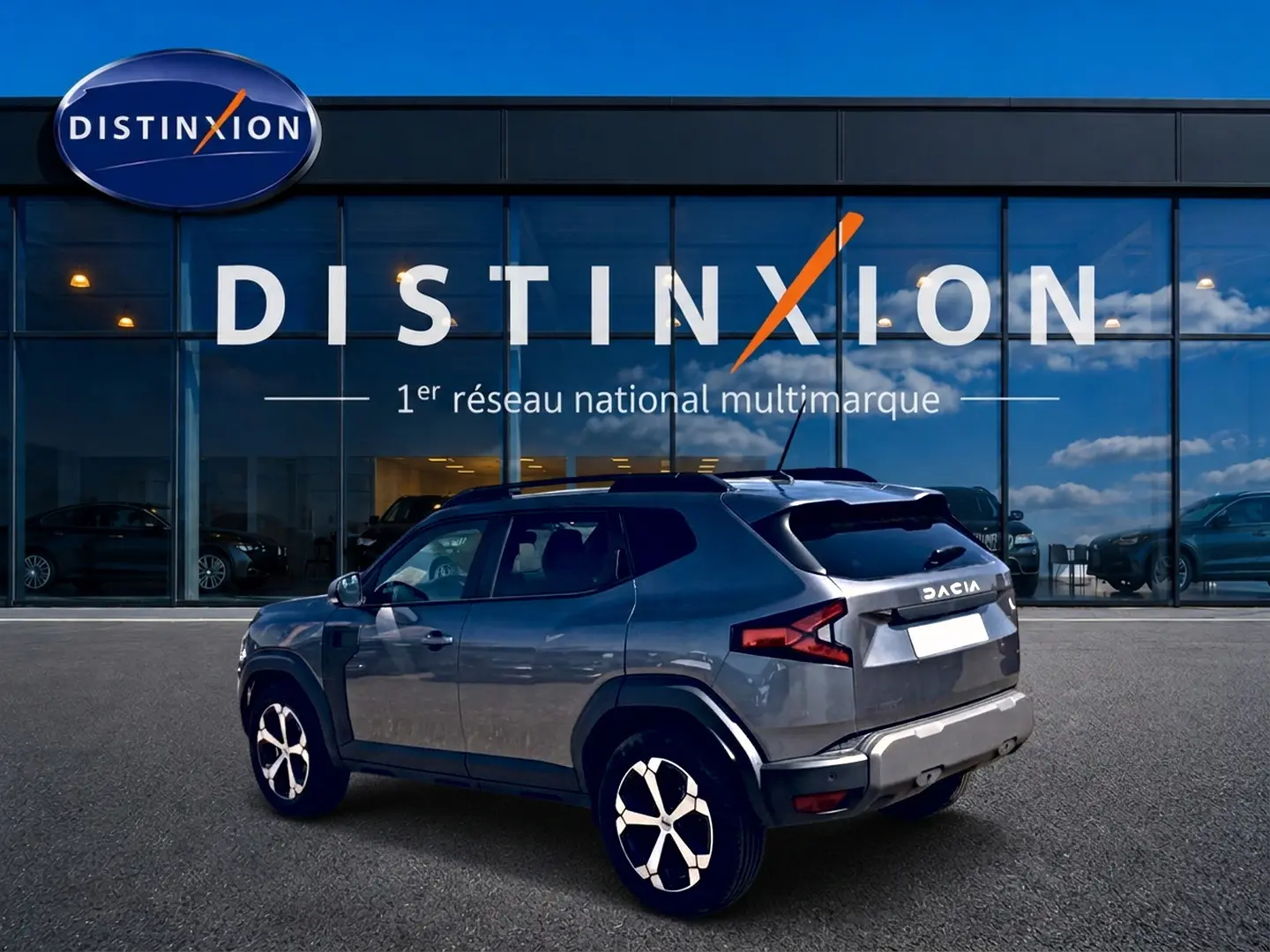 Vue 3/4 arrière droite du Dacia Duster gris schiste 2026 devant une vitrine Distinxion, avec jantes alu et barres de toit.