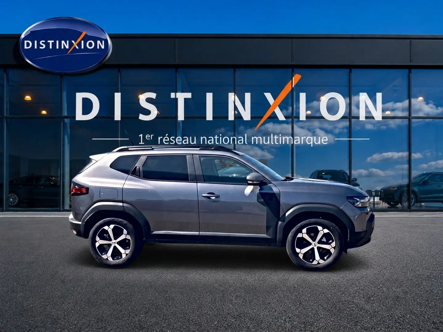 Profil droit du Dacia Duster gris schiste 2026 avec jantes alu et barres de toit devant vitrine Distinxion.