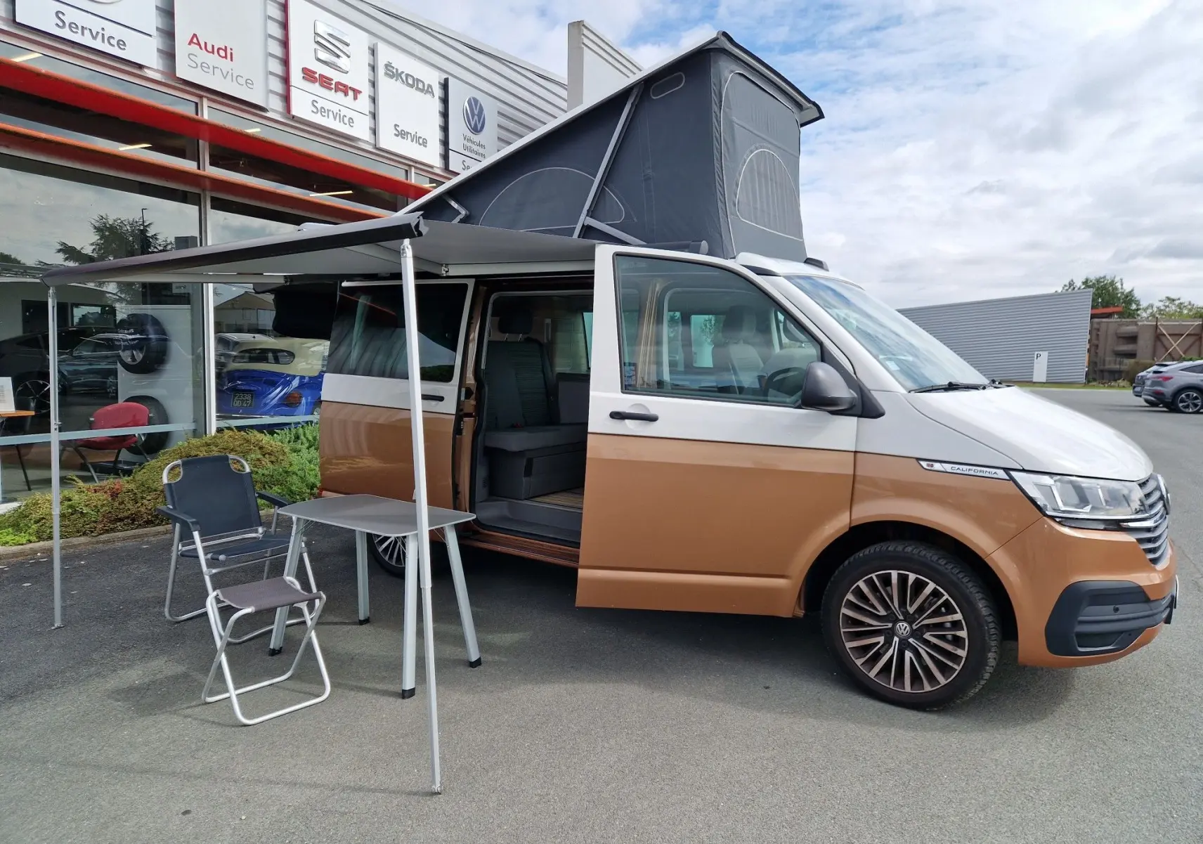 Volkswagen California Cooper bronze 2021 vue côté droit avec toit relevé et auvent déployé sur parking.