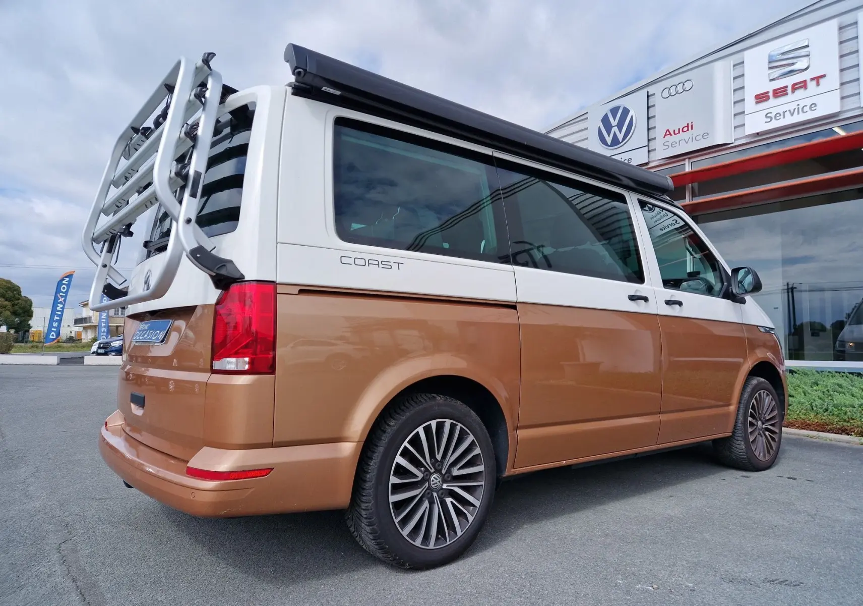 Vue 3/4 arrière droite du Volkswagen California Coast 2021 bicolore Cooper bronze et blanc avec porte-vélos et store latéral noir.
