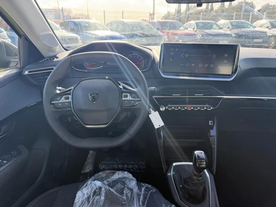 Vue intérieure frontale du poste de conduite d'une Peugeot 208 jaune 2025 avec volant multifonction et écran tactile.