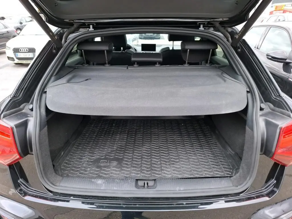 Coffre ouvert vu de l'arrière d'un Audi Q2 noir avec cache-bagages et tapis de coffre en caoutchouc.