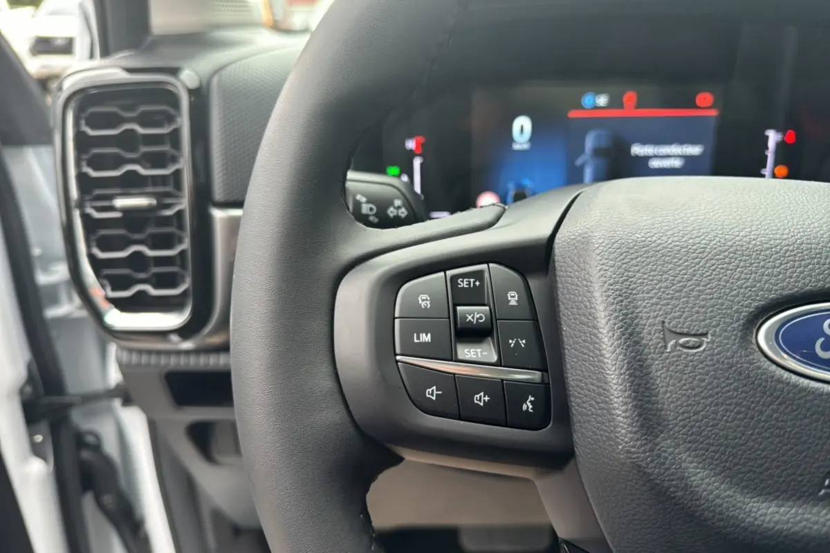 Gros plan sur le volant cuir multifonctions du Ford Ranger blanc glacier, avec commandes intégrées et tableau de bord numérique flou.