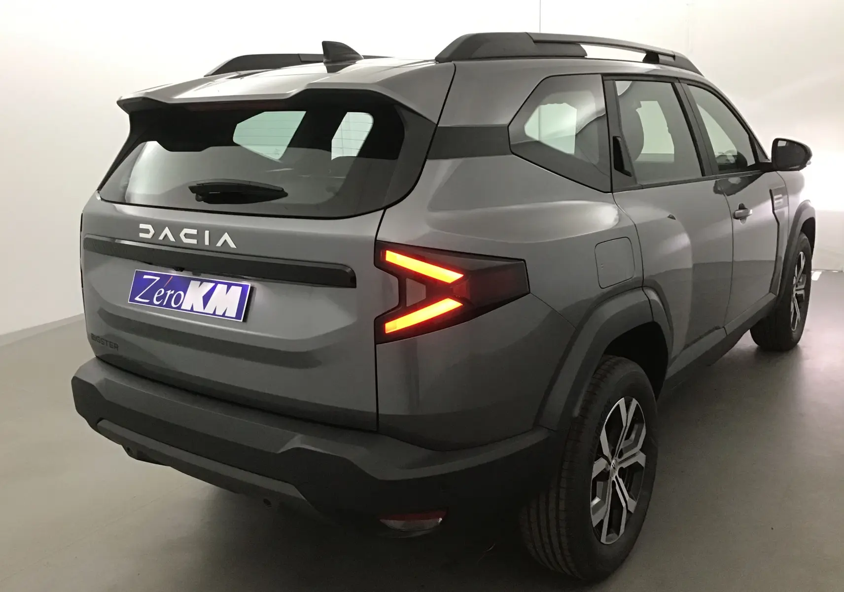 Vue 3/4 arrière droite du Dacia Bigster gris schiste avec feux arrière allumés et logo DACIA visible sur le hayon.
