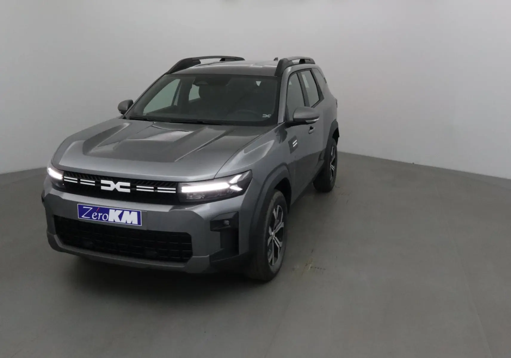 SUV Dacia Bigster gris schiste vu de 3/4 avant droit avec calandre lumineuse et barres de toit noires.