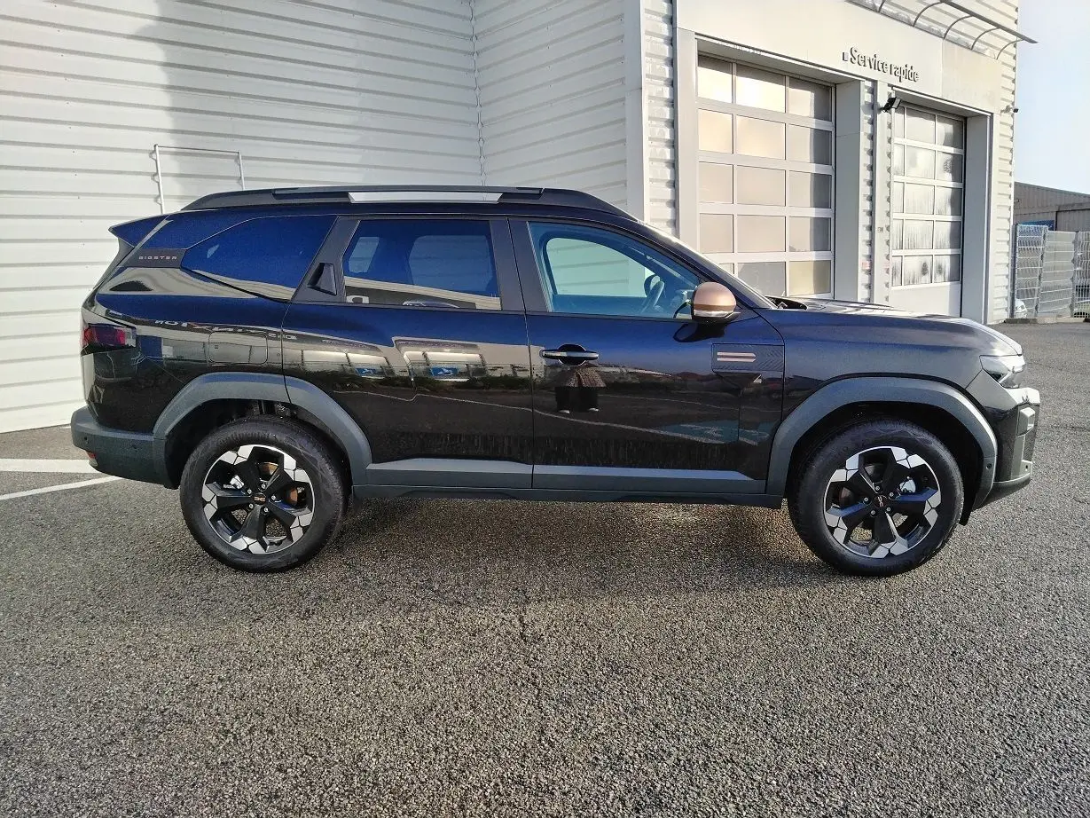 Profil droit d'un Dacia Bigster noir 2025 avec jantes bicolores et rétroviseurs cuivrés, stationné devant un bâtiment blanc.