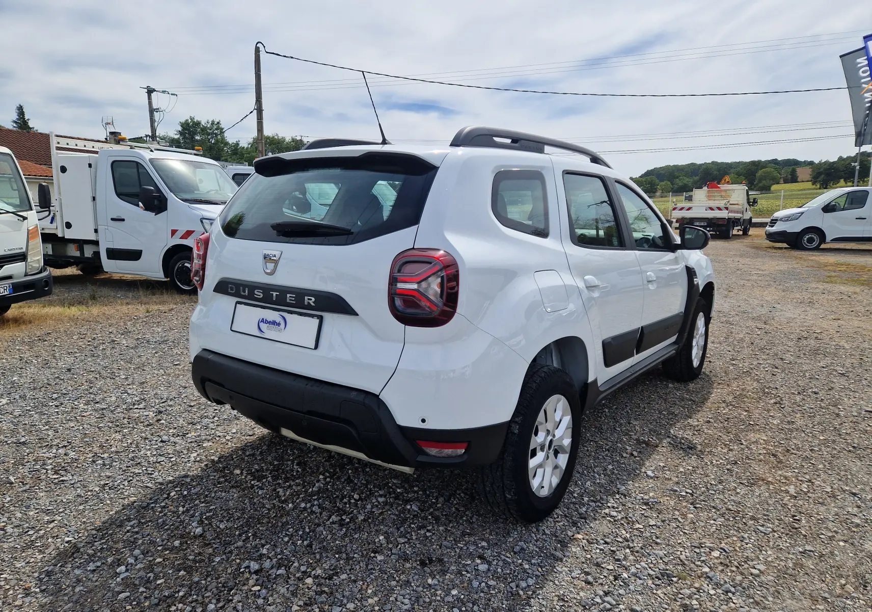 Vue 3/4 arrière droite d'un Dacia Duster blanc 4x4 avec barres de toit et protections latérales noires.