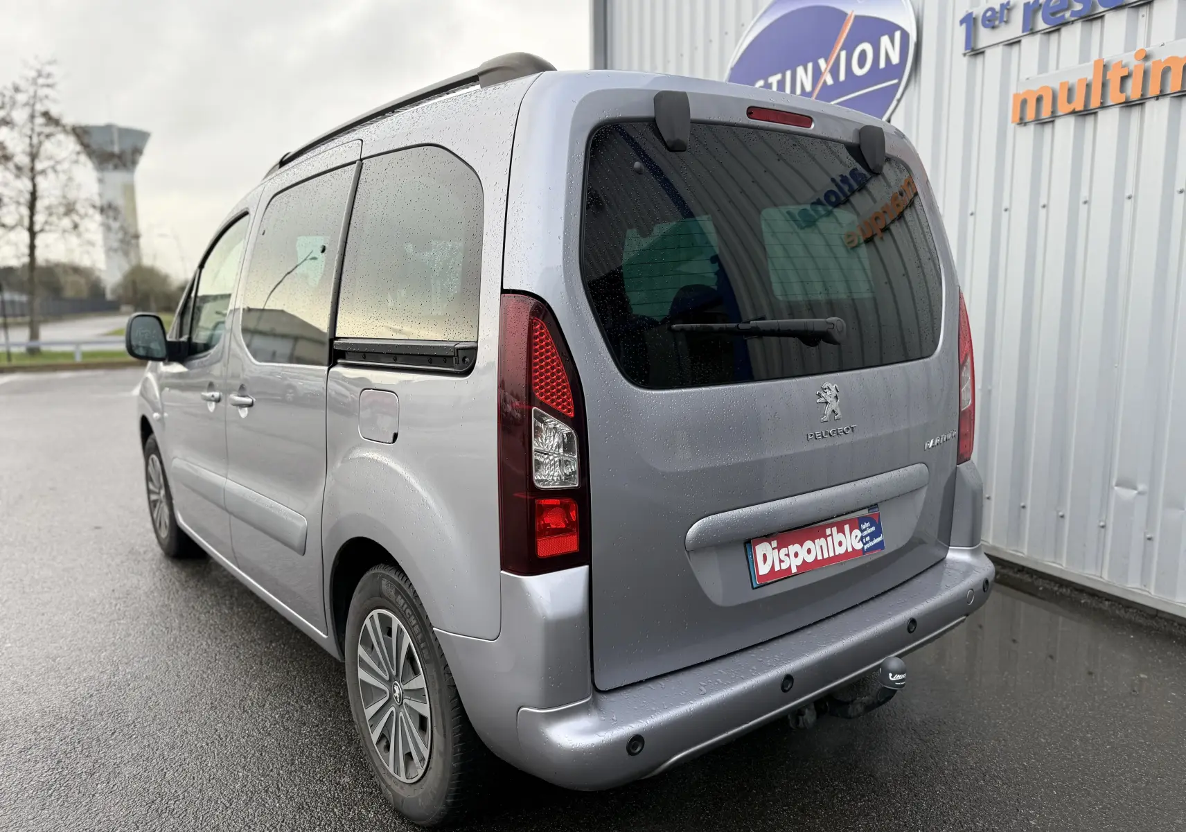 Vue 3/4 arrière droite d’un Peugeot Partner Tepee gris 2018 avec caméra de recul et attelage sous la pluie.