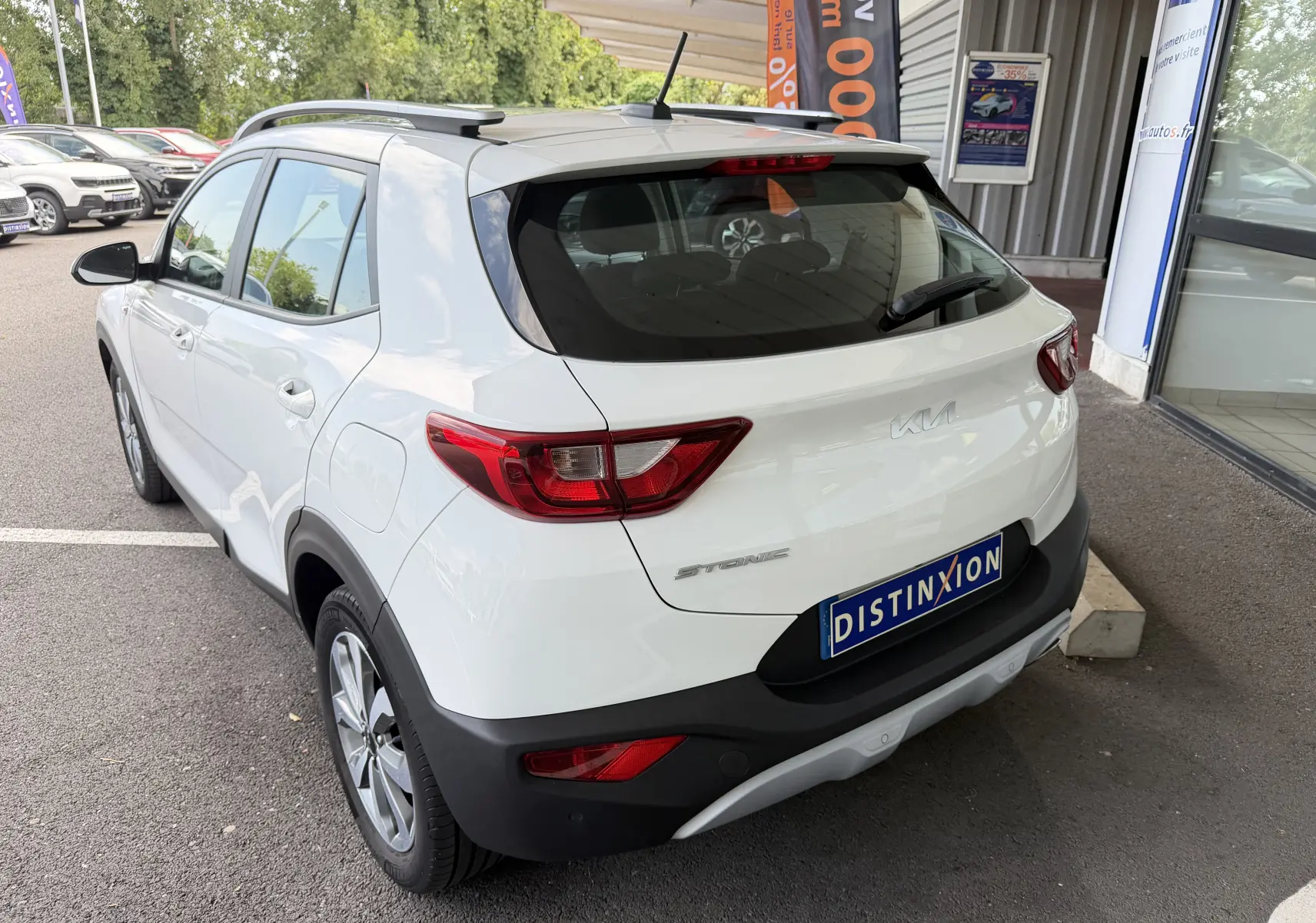 Vue 3/4 arrière droite d’un KIA STONIC blanc 2023 avec toit noir et barres de toit, garé devant un bâtiment commercial.