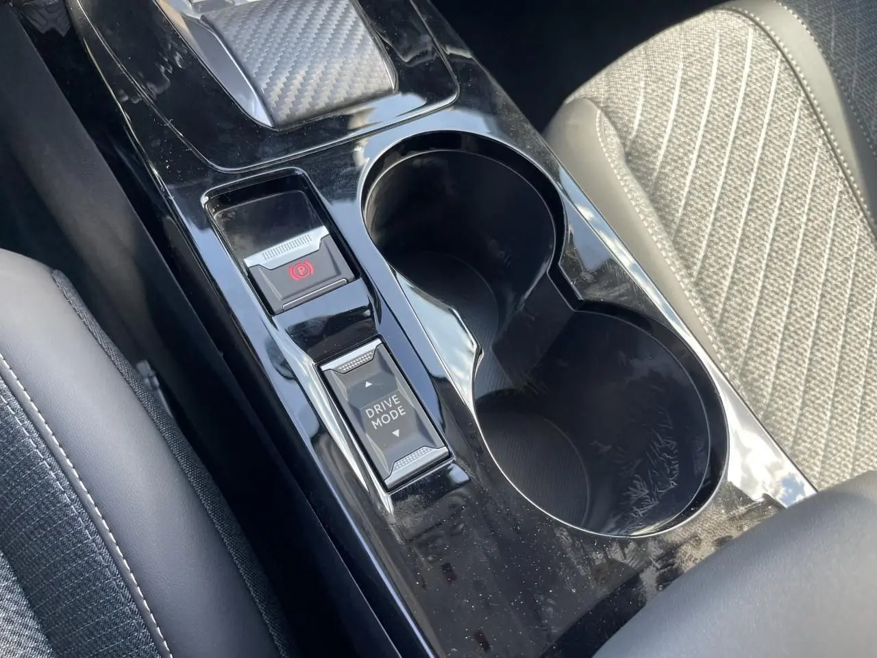 Gros plan sur la console centrale noire brillante avec boutons frein à main électrique et mode de conduite, siège tissu gris à côté.