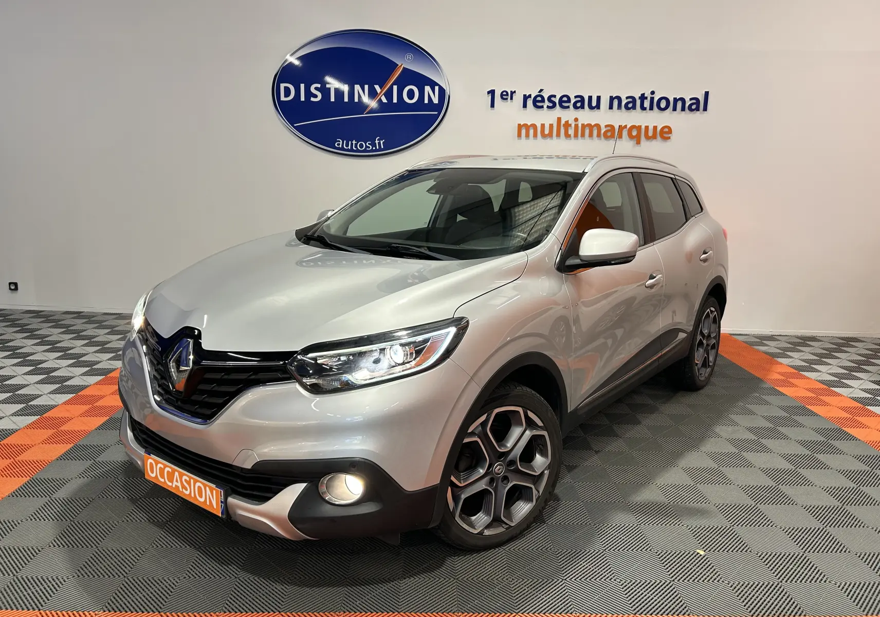 Renault Kadjar gris en 3/4 avant droit, phares et antibrouillards allumés, jantes alliage distinctives visibles.