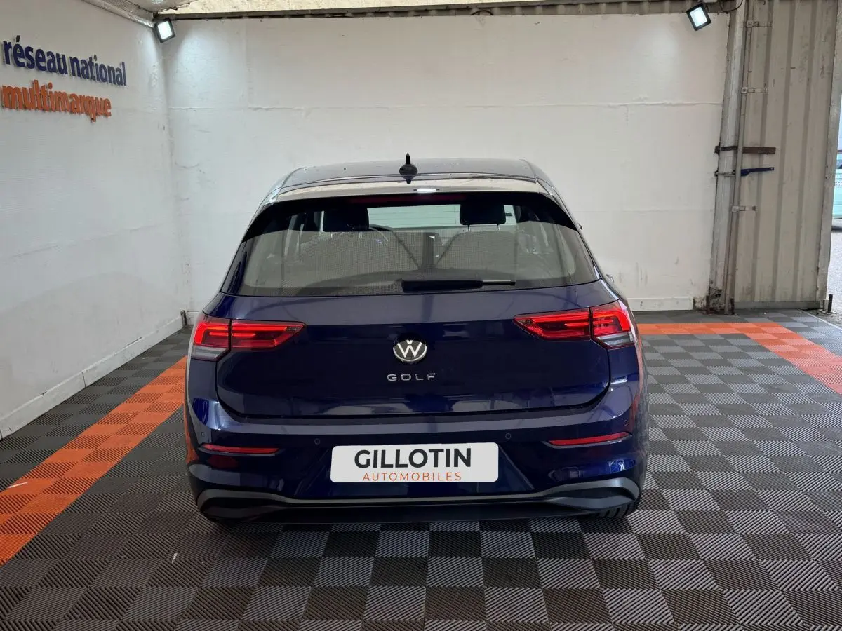 Vue arrière d'une Volkswagen Golf bleu foncé garée en intérieur sur sol à damiers gris et orange.