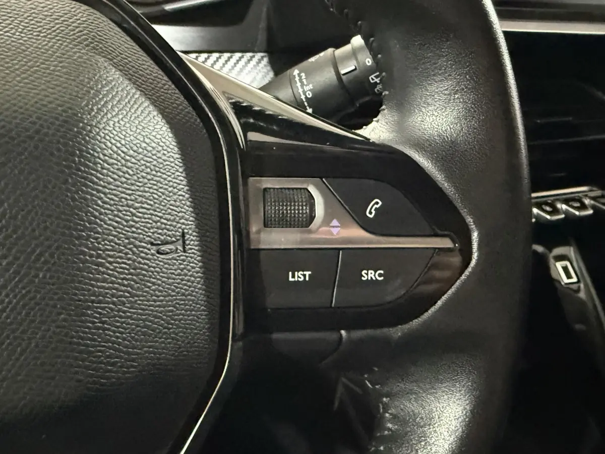 Gros plan sur les commandes gauche du volant noir de la Peugeot 208 1.5 BlueHDi 100 S&S Premium, avec boutons LIST et SRC.