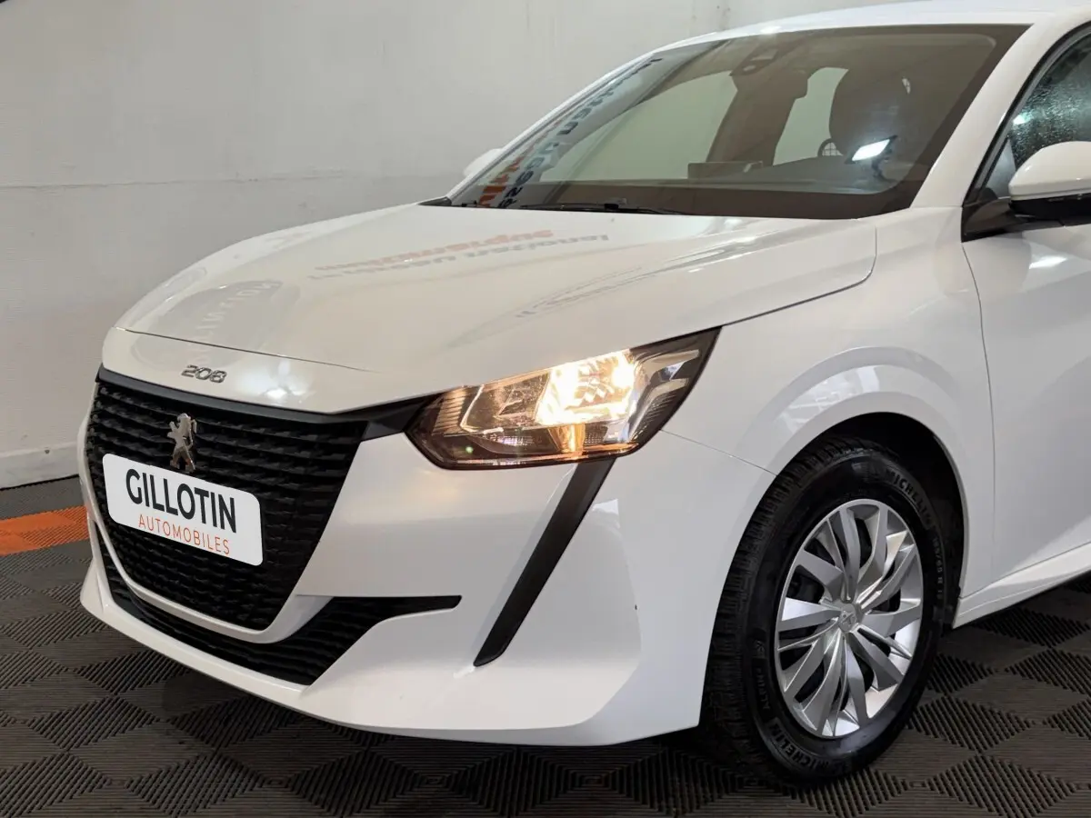 Vue 3/4 avant droite d'une Peugeot 208 blanche avec phares allumés et calandre noire distinctive.