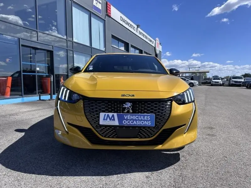 Vue avant d'une Peugeot 208 jaune faro 2020 GT line avec calandre noire et feux LED distinctifs, stationnée devant un garage.