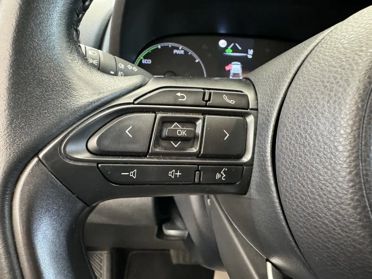 Gros plan sur les commandes multifonctions du volant cuir noir de la Toyota Yaris hybride blanche, tableau de bord en arrière-plan.