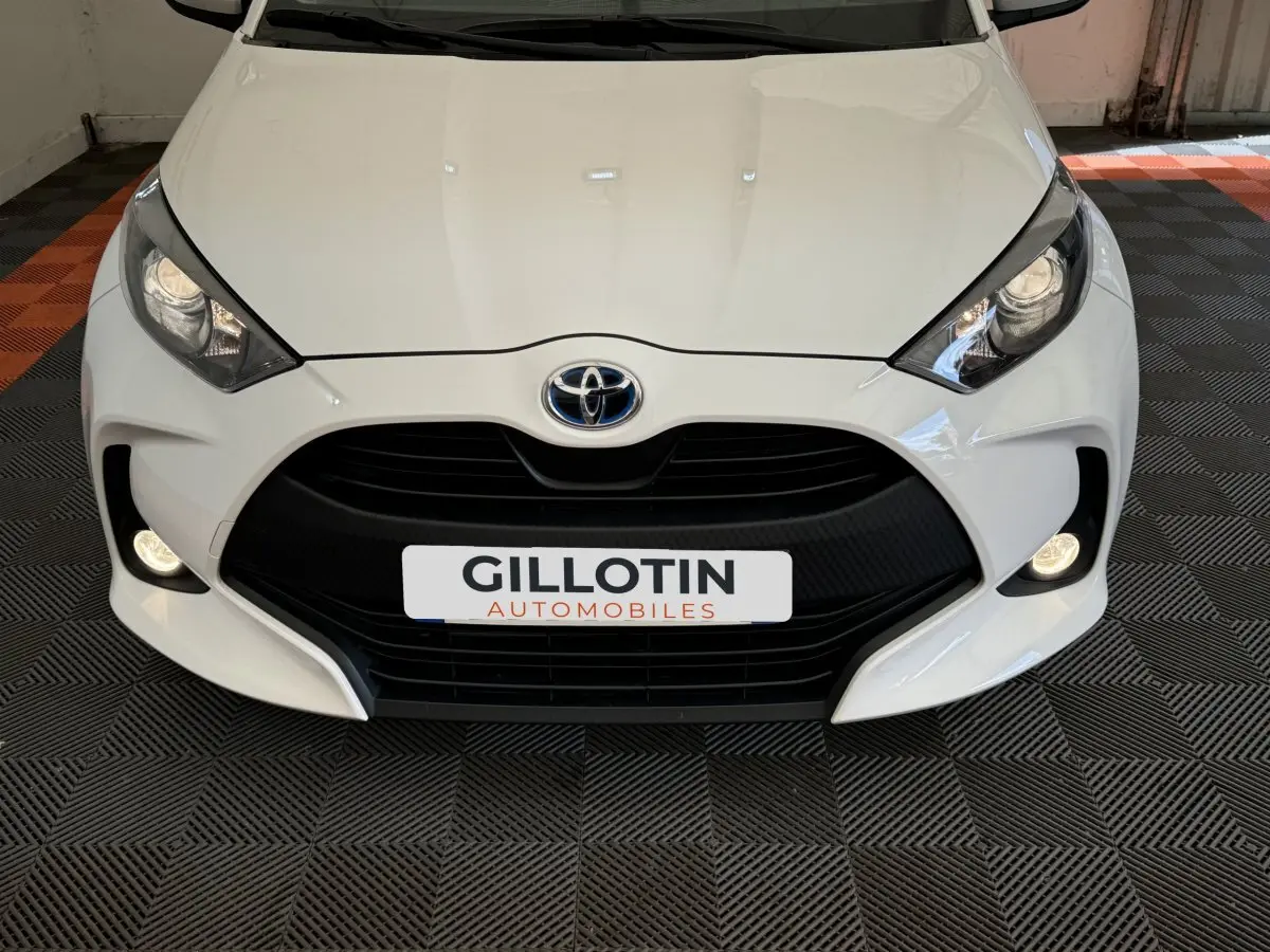 Vue frontale rapprochée d'une Toyota Yaris blanche avec calandre noire et phares allumés dans un showroom.