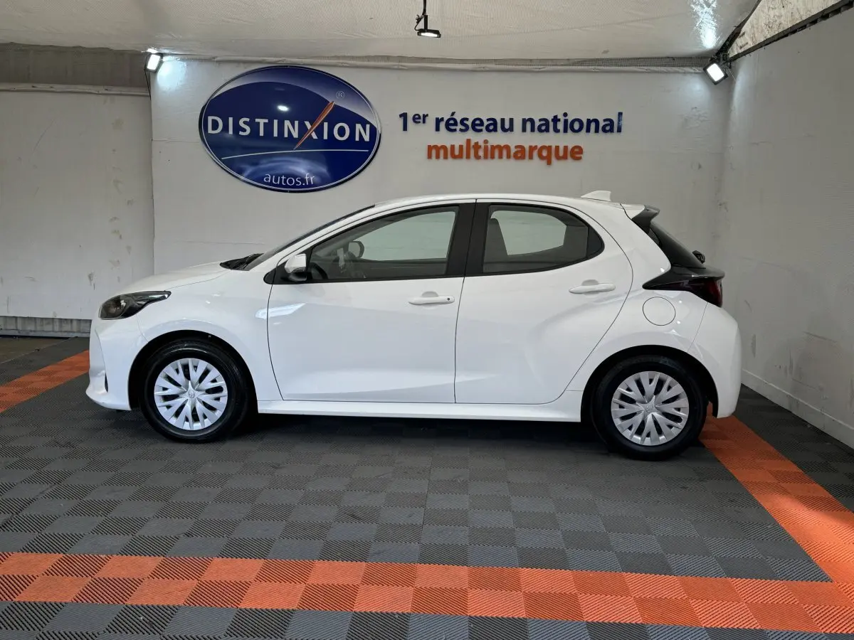 Profil côté gauche d'une Toyota Yaris hybride blanche 2022, dans un showroom avec sol gris et bandes orange.