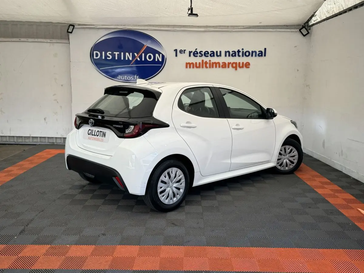 Toyota Yaris hybride blanche vue 3/4 arrière droit en intérieur, avec toit noir et jantes standard.