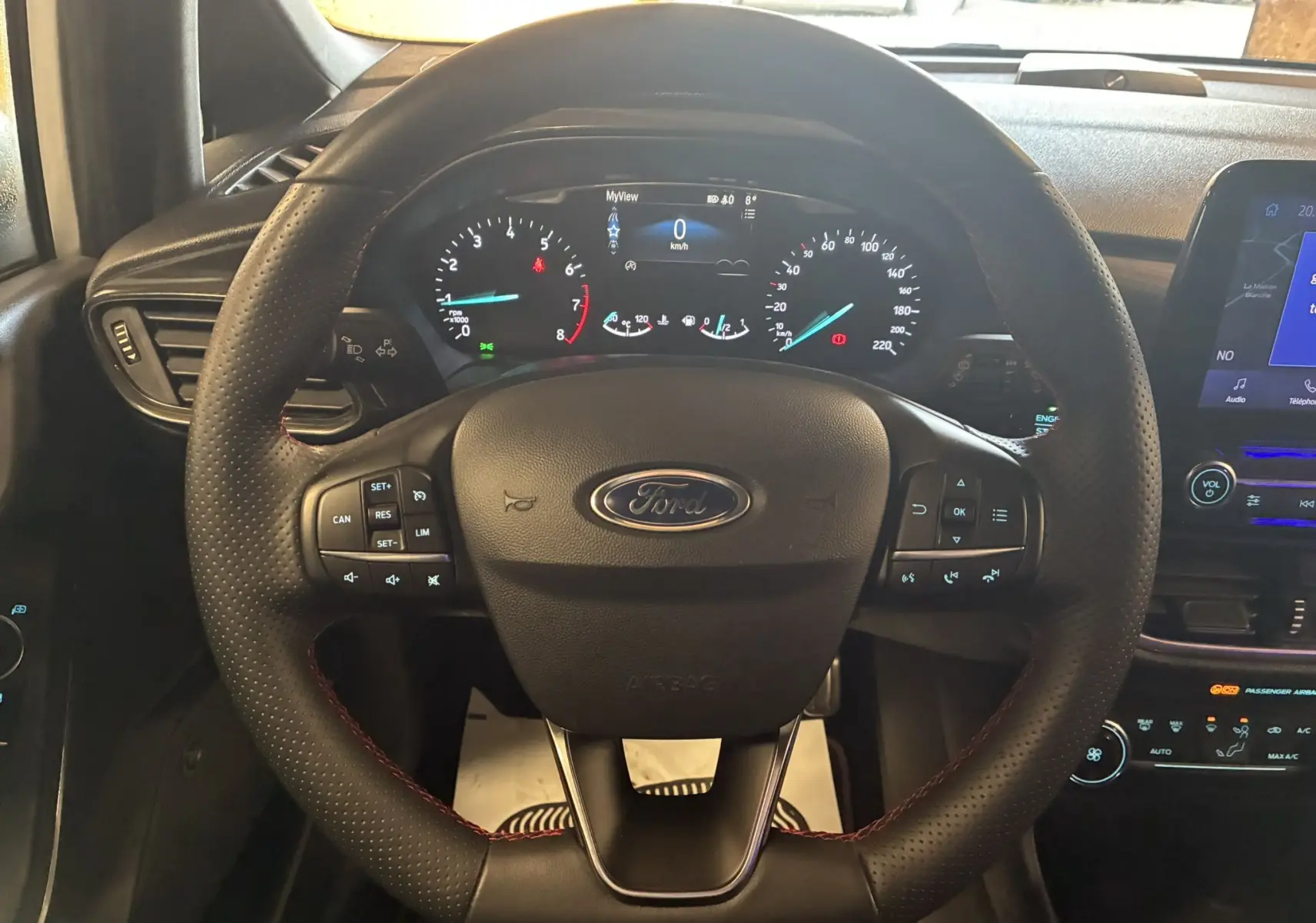 Vue rapprochée du volant en cuir perforé avec surpiqûres rouges de la Ford Fiesta 1.0 EcoBoost 95 ST Line, tableau de bord numérique allumé.