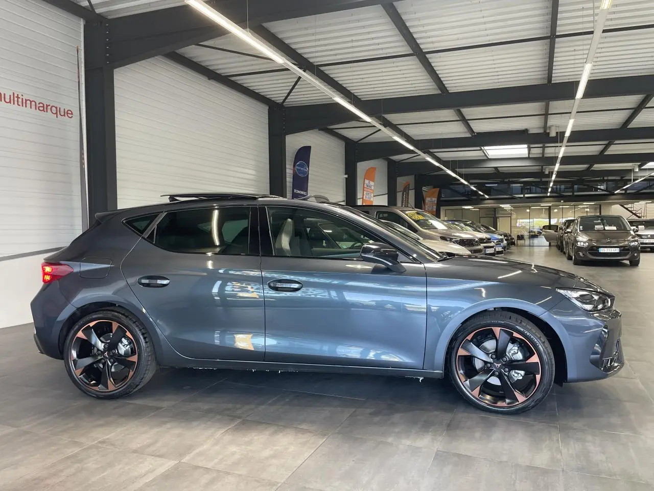 Profil droit d'une CUPRA Leon 2.0 TDI gris magnétique avec jantes alliage 18'' cuivrées en showroom.