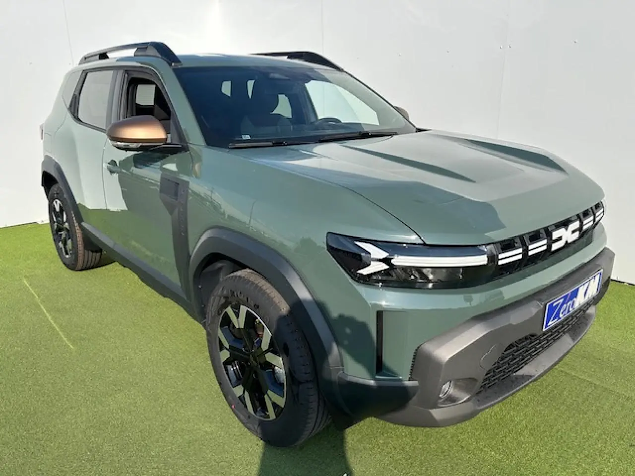 Dacia Duster III kaki dusty en 3/4 avant droit, avec jantes alliage noires et barres de toit longitudinales noires.