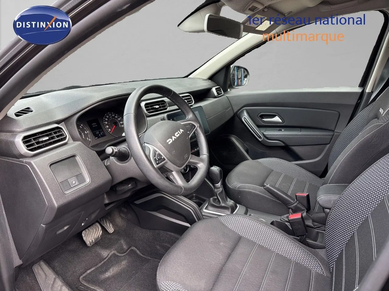 Vue intérieure côté conducteur du Dacia Duster 2024 noir, montrant le tableau de bord et les sièges en tissu noir.