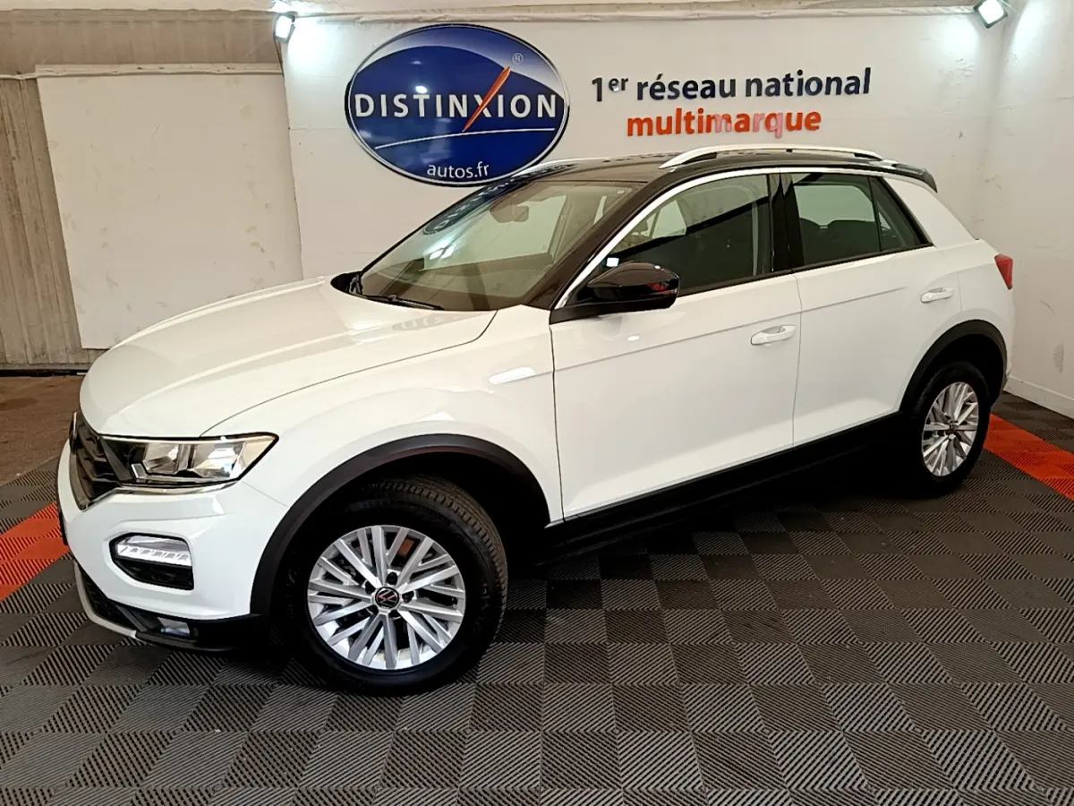 Vue 3/4 avant droite du Volkswagen T-ROC blanc avec jantes alliage et barres de toit noires en intérieur showroom.