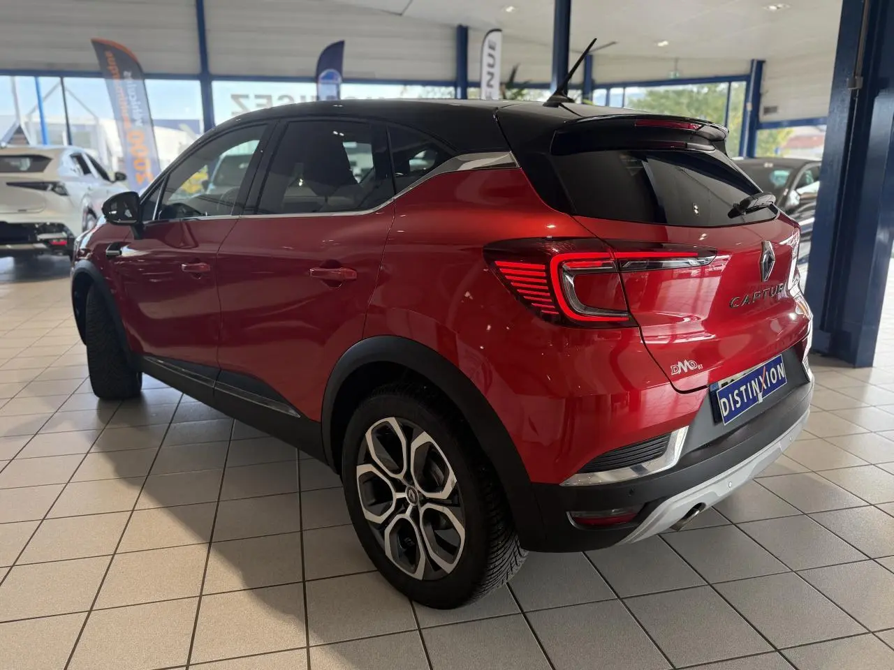 Vue 3/4 arrière droite d'un Renault Captur rouge foncé avec toit noir en showroom, soulignant ses feux arrière LED et jantes stylées.