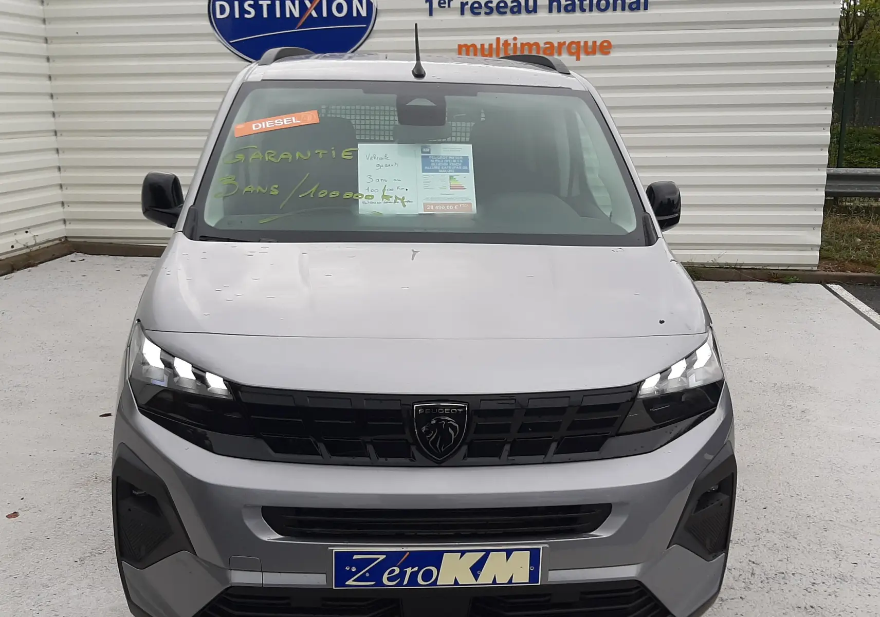 Vue avant d'un Peugeot Rifter gris acier 2026 avec calandre noire et plaques "Zéro KM" sur un parking.