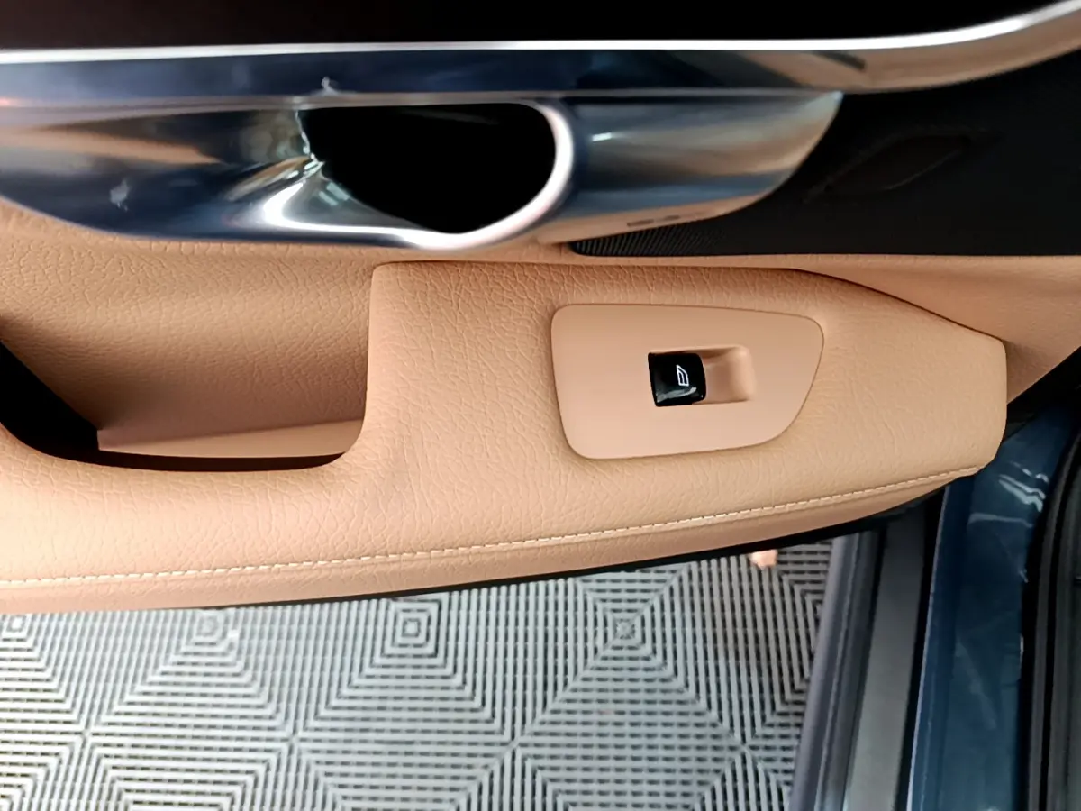 Détail de la commande de lève-vitre sur la porte côté conducteur en cuir beige, avec poignée métal et bord bleu extérieur visible.