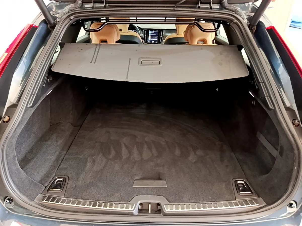 Coffre ouvert vu de l'arrière d'un Volvo V90 Cross Country bleu, avec cache-bagages et intérieur beige visible.