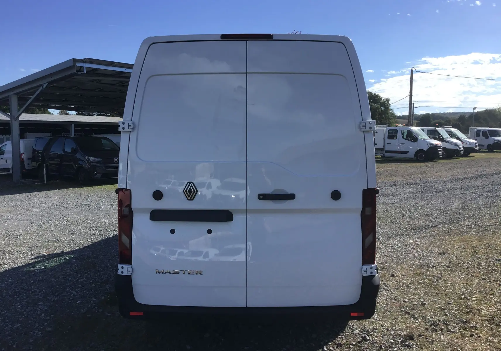 Vue arrière d'un utilitaire Renault Master blanc, portes battantes fermées, dans un parking extérieur ensoleillé.