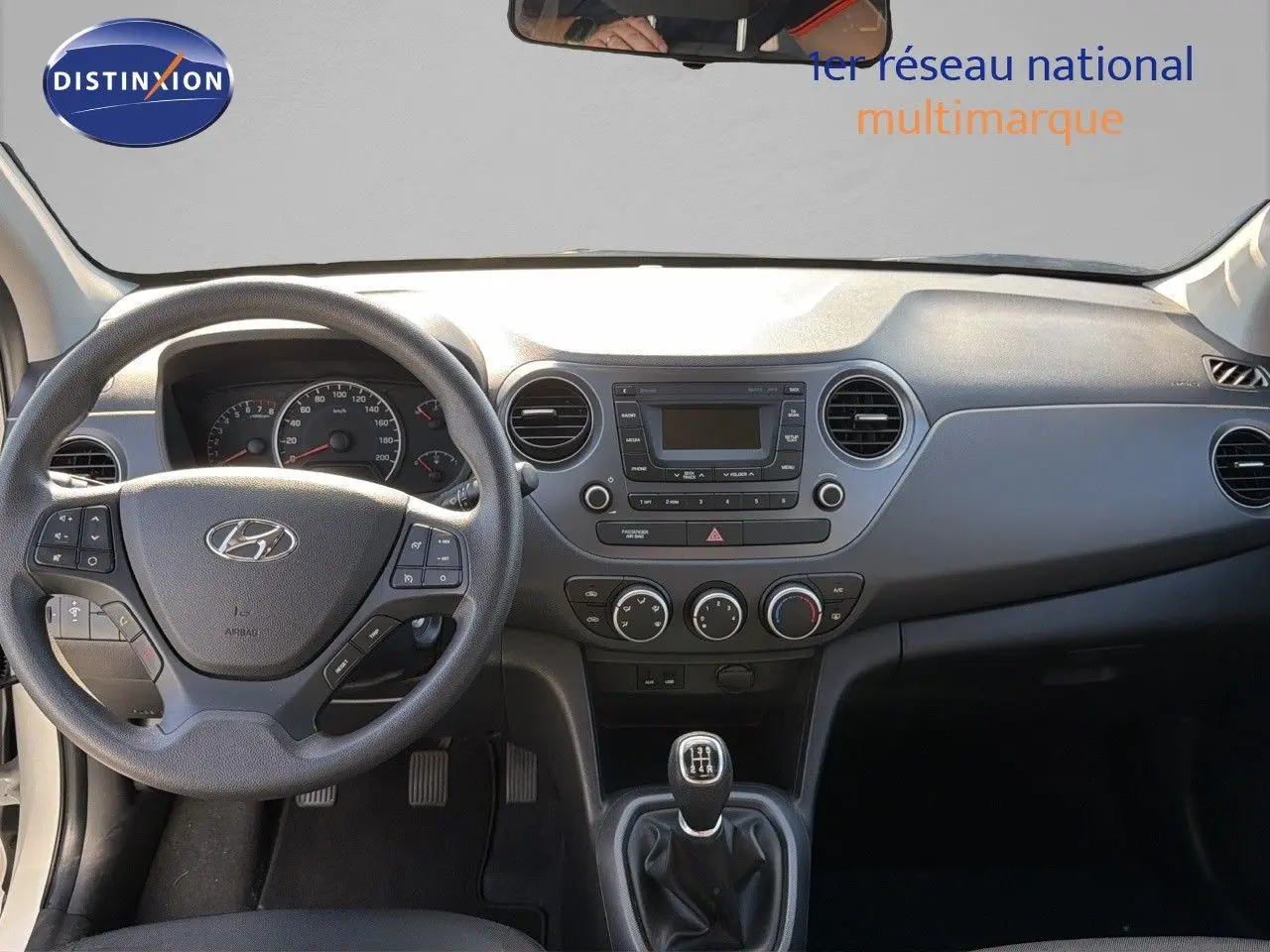 Vue intérieure du tableau de bord et volant noir d'une Hyundai i10 blanche 2018 avec boîte manuelle et commandes au volant.