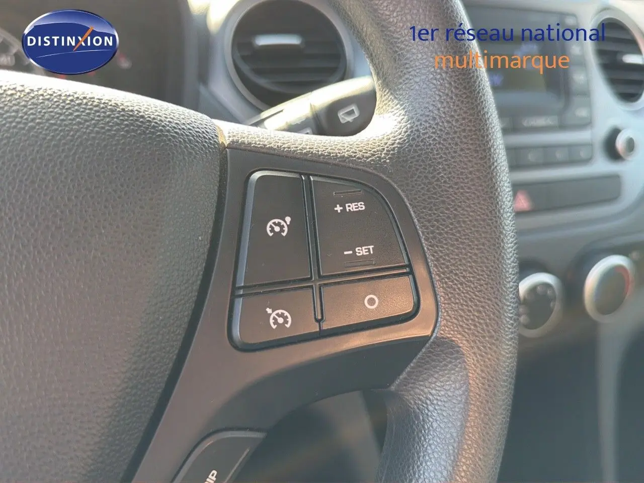 Gros plan sur les commandes de régulateur de vitesse au volant d'une Hyundai i10 blanche, intérieur visible en arrière-plan.