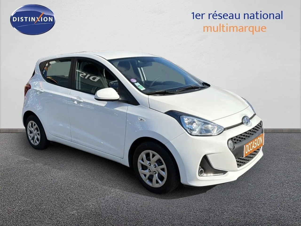 Hyundai i10 blanche vue 3/4 avant droit, compacte cinq portes avec jantes alliage et calandre noire distinctive.