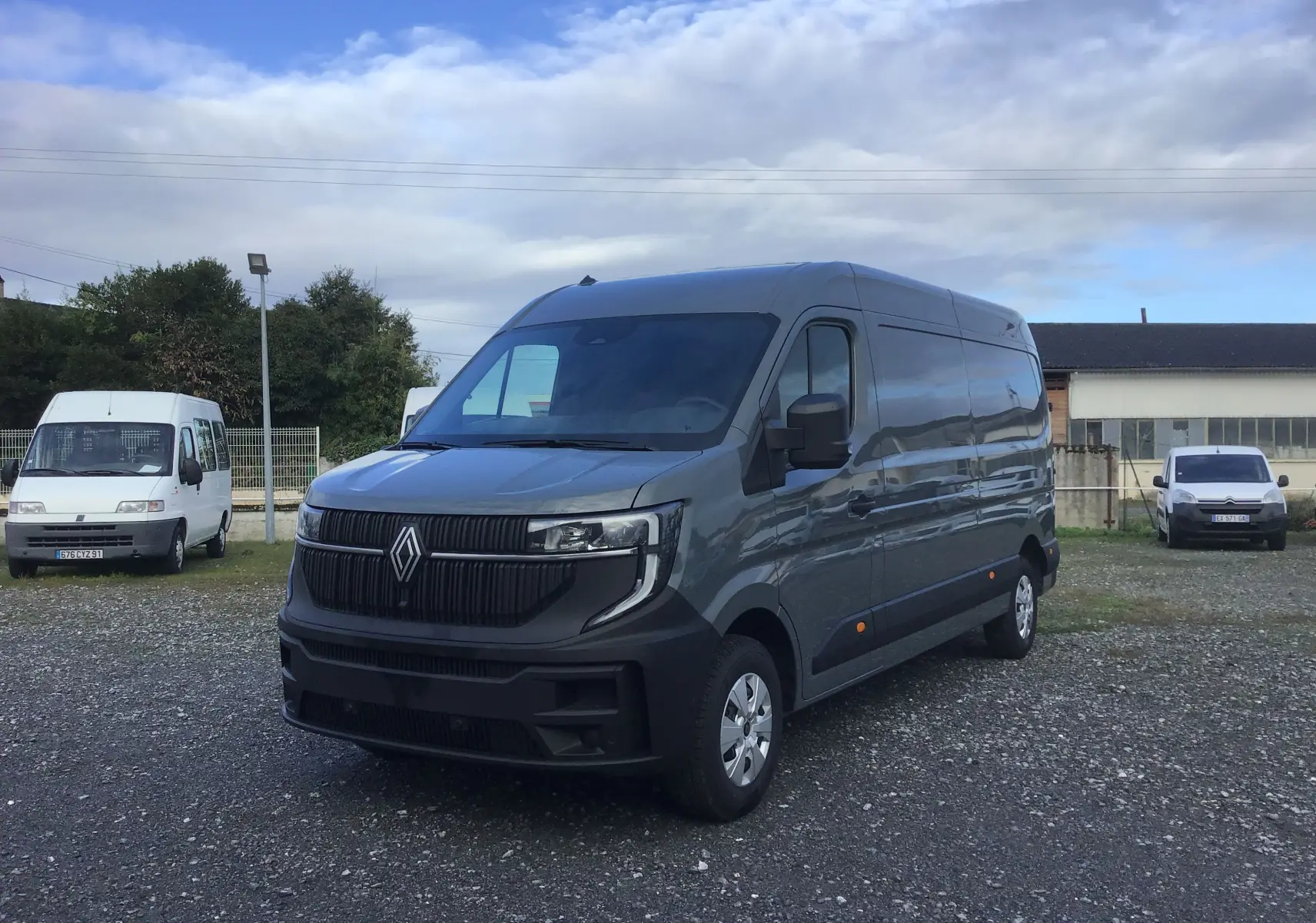 Renault Master gris urbain vu en 3/4 avant droit sur un sol gravillonné avec bâtiments en arrière-plan.