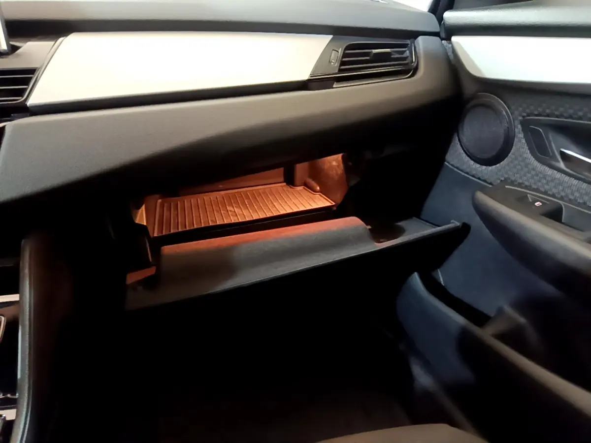 Intérieur côté passager de la BMW Série 2 Gran Tourer 218d, boîte à gants ouverte avec éclairage ambre et garnitures noires.