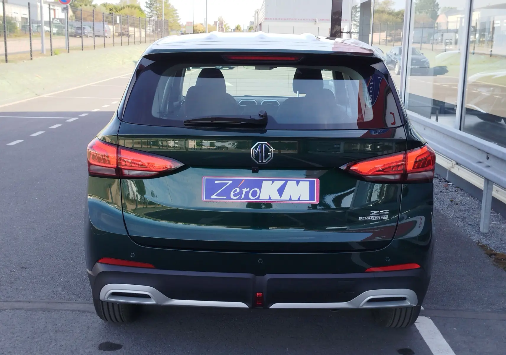 Vue arrière d'un MG Motor ZS Emerald Green Metal avec feux allumés et badge hybride visible sur un parking.