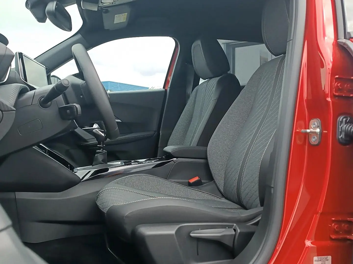 Vue intérieure côté conducteur de la Peugeot 2008 rouge elixir, montrant les sièges en tissu gris et la console centrale.