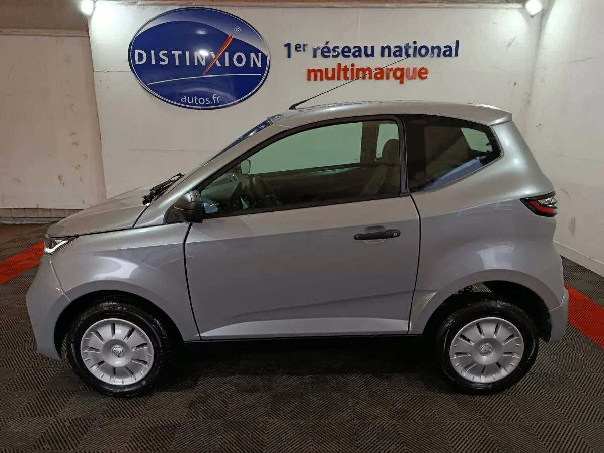 Profil côté gauche d'une Aixam Ambition 0.5 City Pack gris clair 2026 avec rétroviseur caméra conducteur en intérieur showroom.
